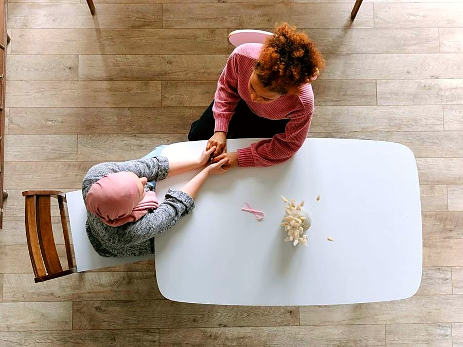 A caregiver holds the hands of an oncology patient wearing a headscarf, offering support at home
